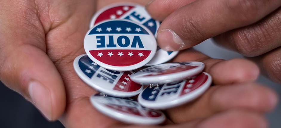 Hands holding round lapel pins that say VOTE